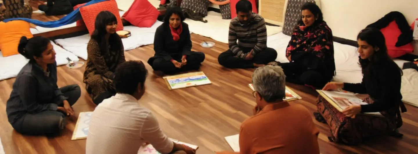 Seven people seated in a circle on a wooden floor with papers, participating in a group activity.