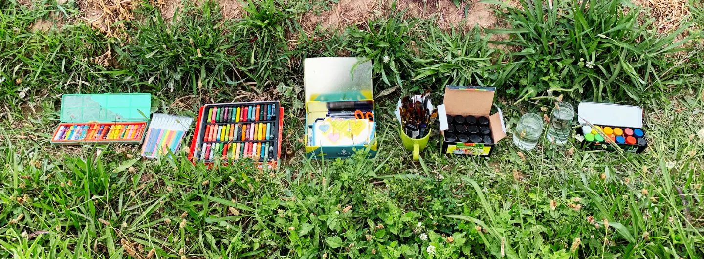 Art supplies including crayons, pastels, brushes, paint jars, water cups, and a watercolor set on green grass.