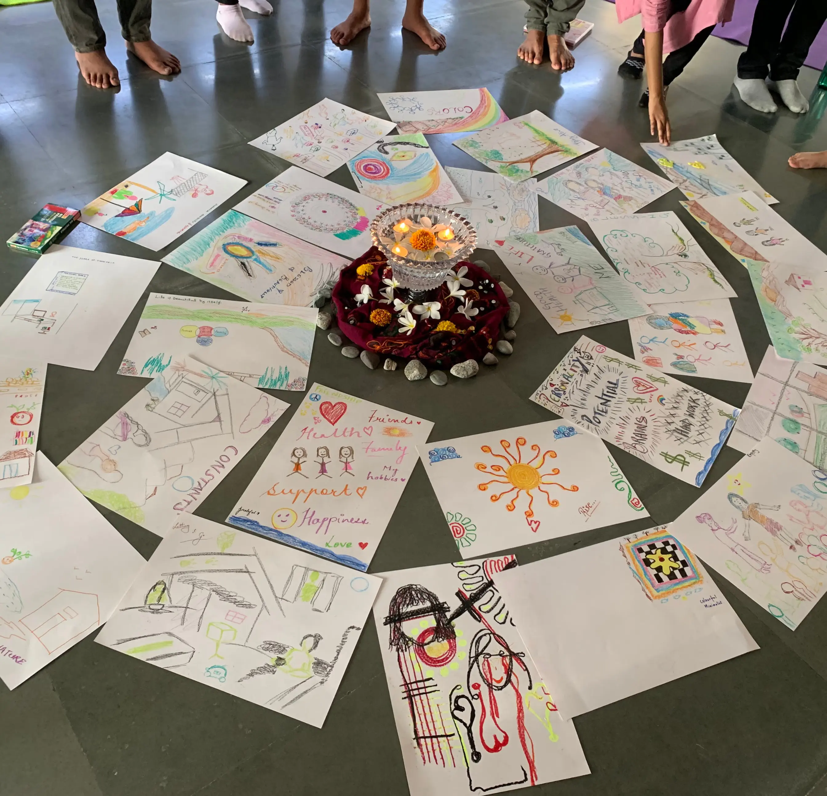 A group of drawings arranged in a circle on the floor surrounds a central bowl with floating candles and flowers.
