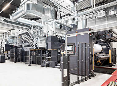 Industrial printing machines with control panels and large paper rolls in a spacious factory.