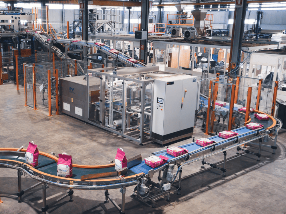 Factory conveyor belt transporting pink and white packaged goods around large machinery in a warehouse.
