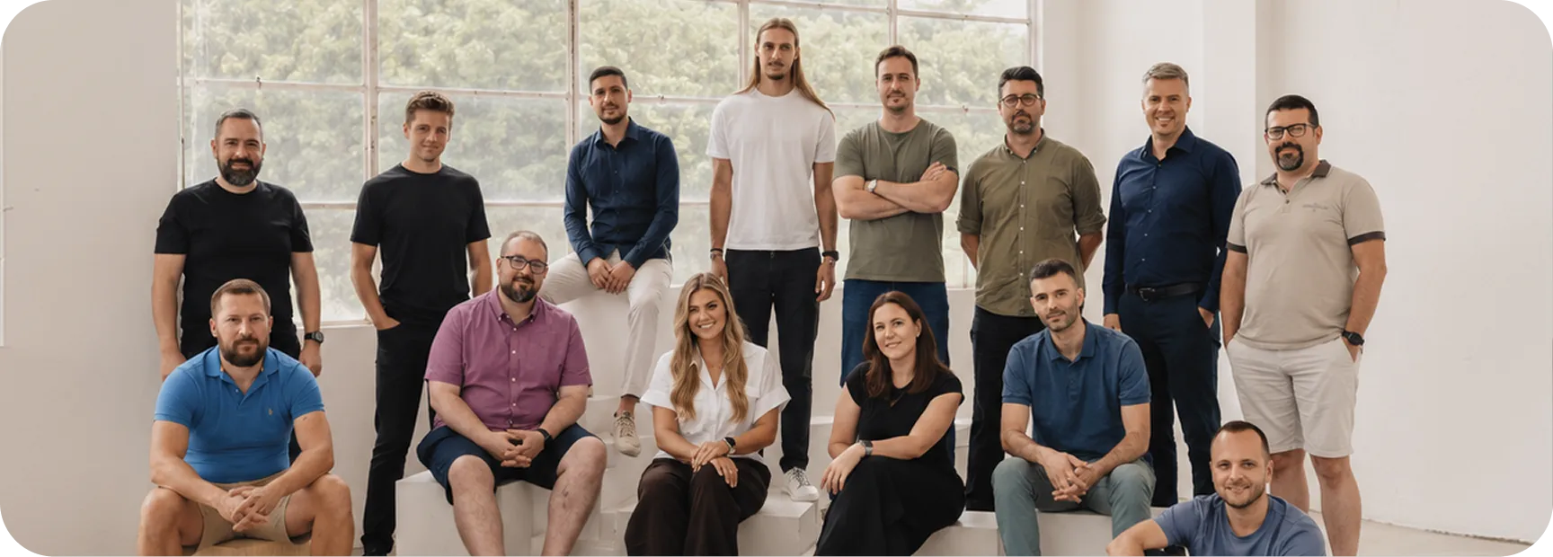 Group of 15 adults posing indoors against a large window, some seated and some standing.