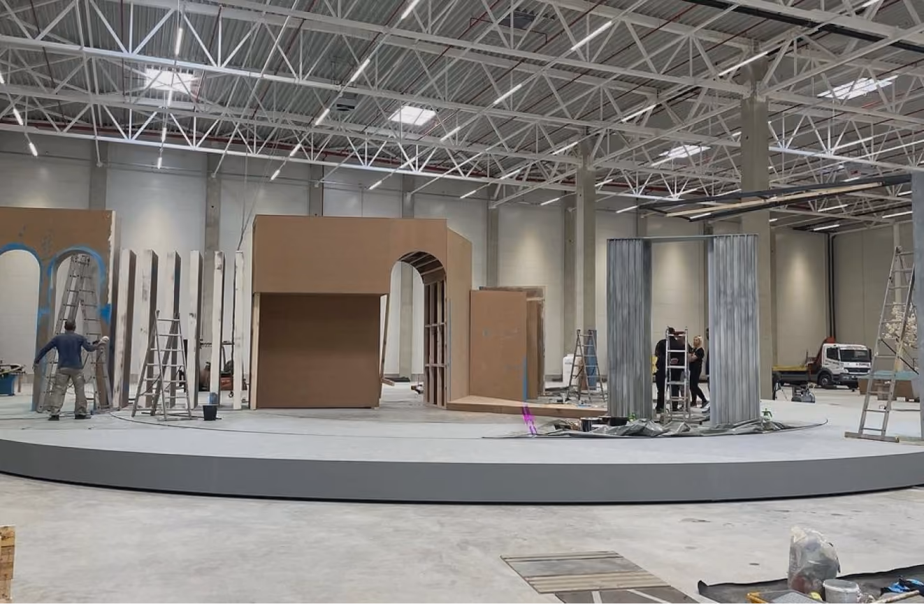 Large indoor space with people assembling a stage set featuring arches, ladders, and panels.
