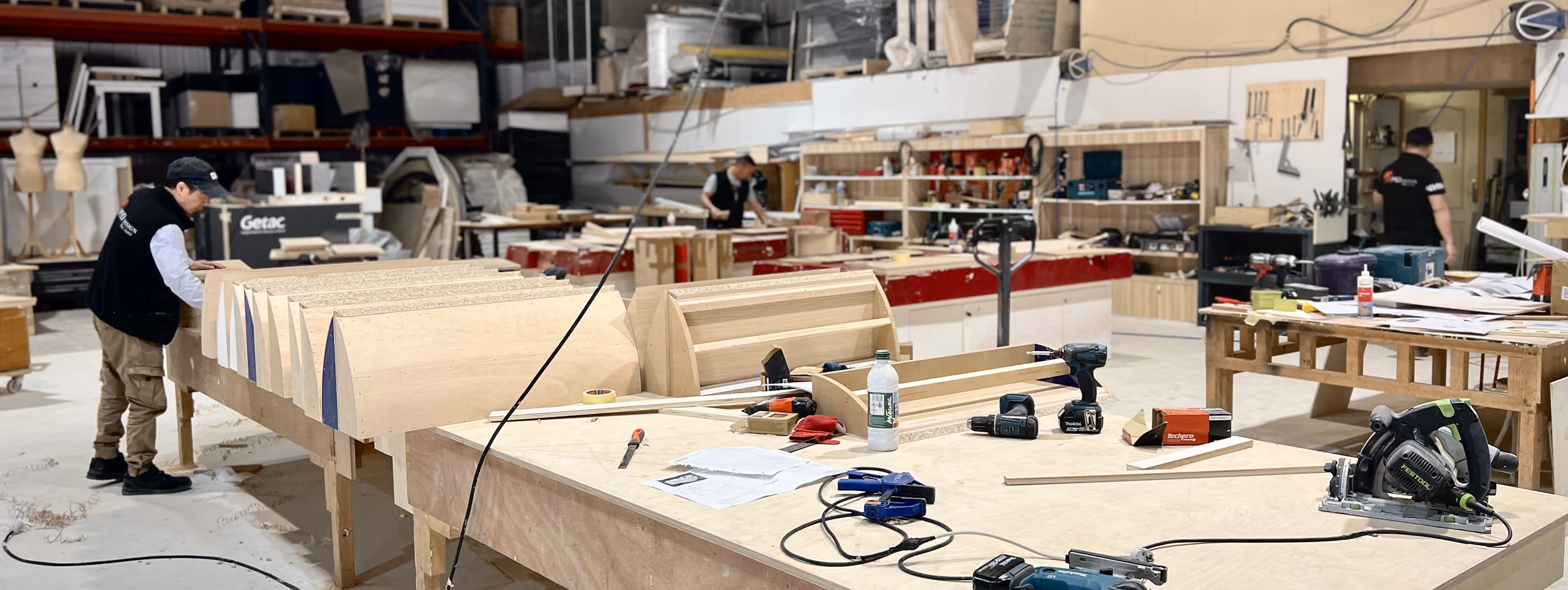 A woodworking shop with tools and wooden parts on workbenches and workers engaged in tasks.