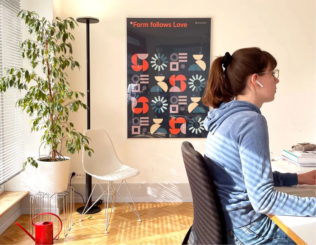 Frau mit Ponytail und Brille arbeitet am Tisch, Zimmer mit Poster, Zimmerpflanze und Stuhl.