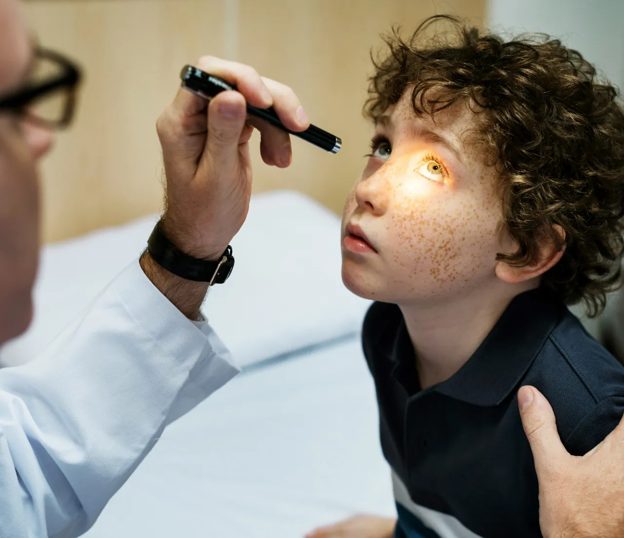 Arzt prüft das Auge eines Kindes mit Locken und Sommersprossen mit einer Taschenlampe.