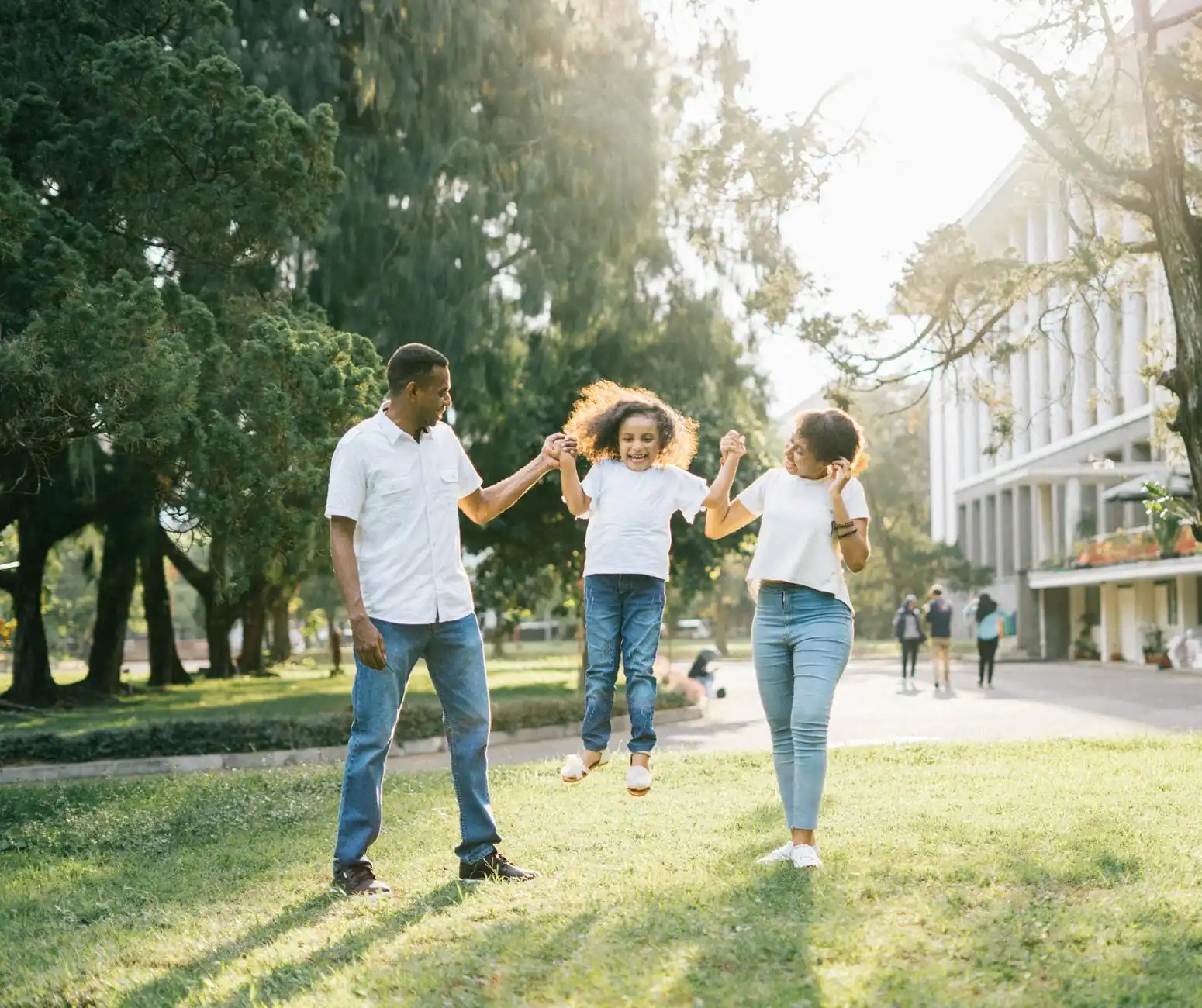 Parents holding hands and swinging their young child between them on a sunny grassy lawn.