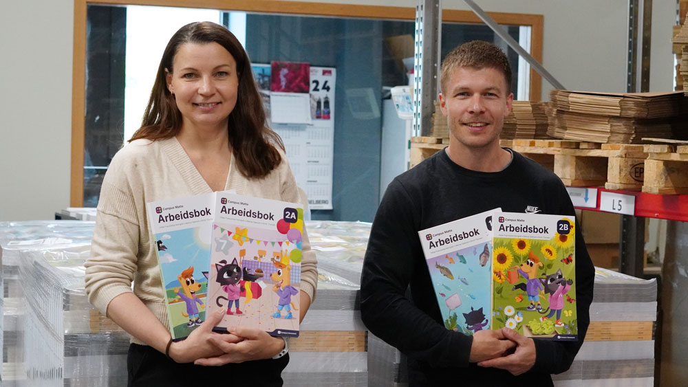 Forfatterne av arbeidsbøkene, Kirsti Thorsen og Marius Saanum, med arbeidsbøker i hendene. De står foran to paller med arbeidsbøker.