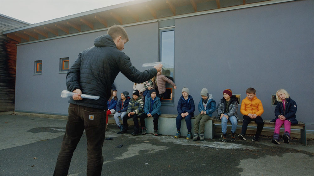 Lærer og en gruppe elever utendørs. Læreren snakker med elevene som sitter på en benk og følger med, fotografi