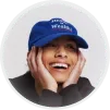 Smiling person wearing a blue hat and resting their head in their hands against a dark background.