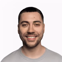 Smiling young man with short dark hair and trimmed facial hair wearing a light gray shirt against a plain white background.