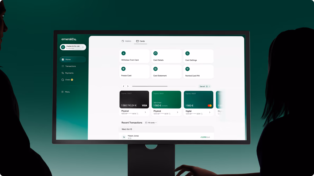 Two people in silhouette viewing a desktop monitor displaying a financial dashboard with various bank card options and recent transactions in an app called emerald24.