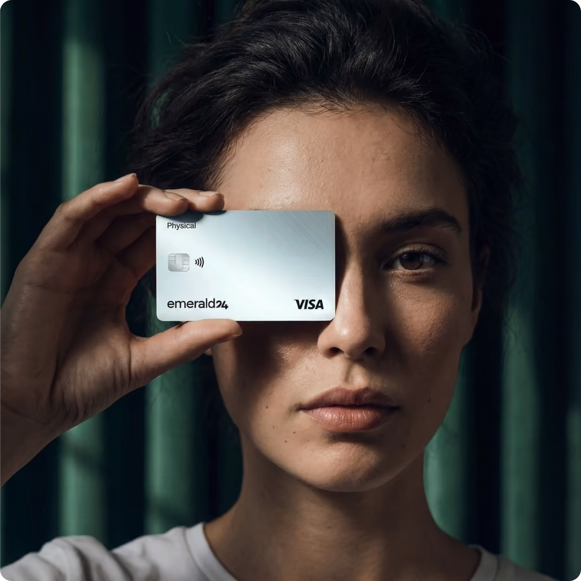 Woman holding a white Visa Emerald24 credit card in front of her right eye against a dark green curtain background.