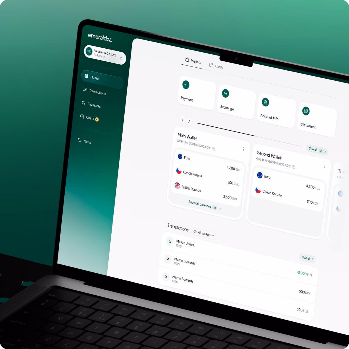 Laptop screen showing Emerald24 digital wallet interface with two wallets displaying balances in Euro, Czech Koruna, and British Pounds, and recent transactions listed below.