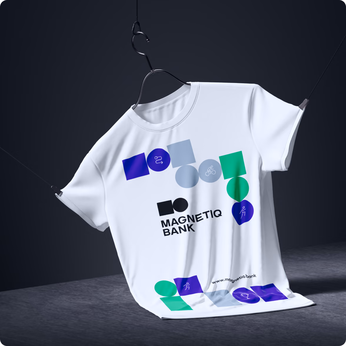 White T-shirt hanging on a wire hanger against a dark background, featuring colorful geometric shapes and icons around the text 'MAGNETIQ BANK' with a website URL at the bottom.