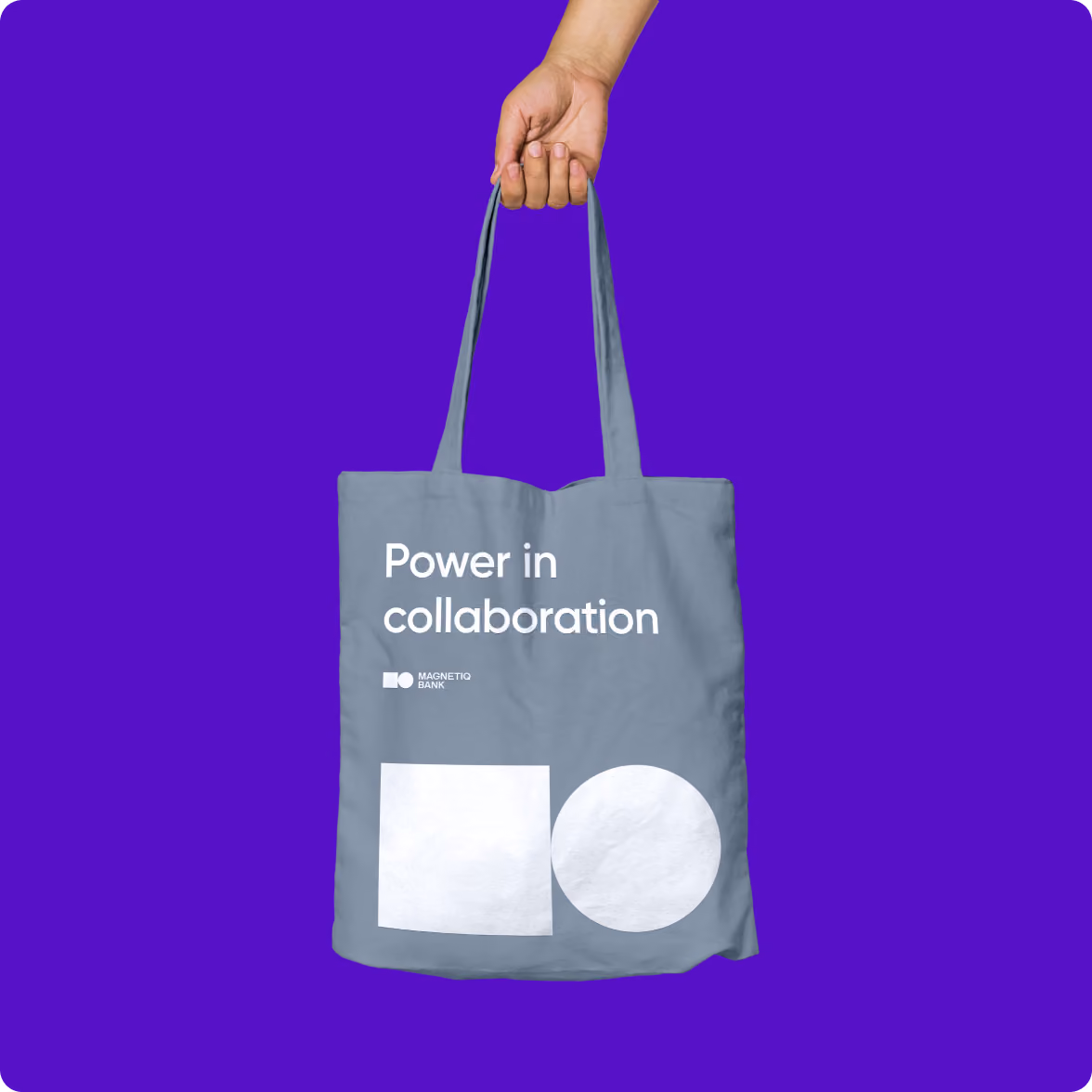 A hand holding a gray tote bag with white text that reads 'Power in collaboration' and 'Magnetiq Bank' above a white square and circle design, set against a solid purple background.