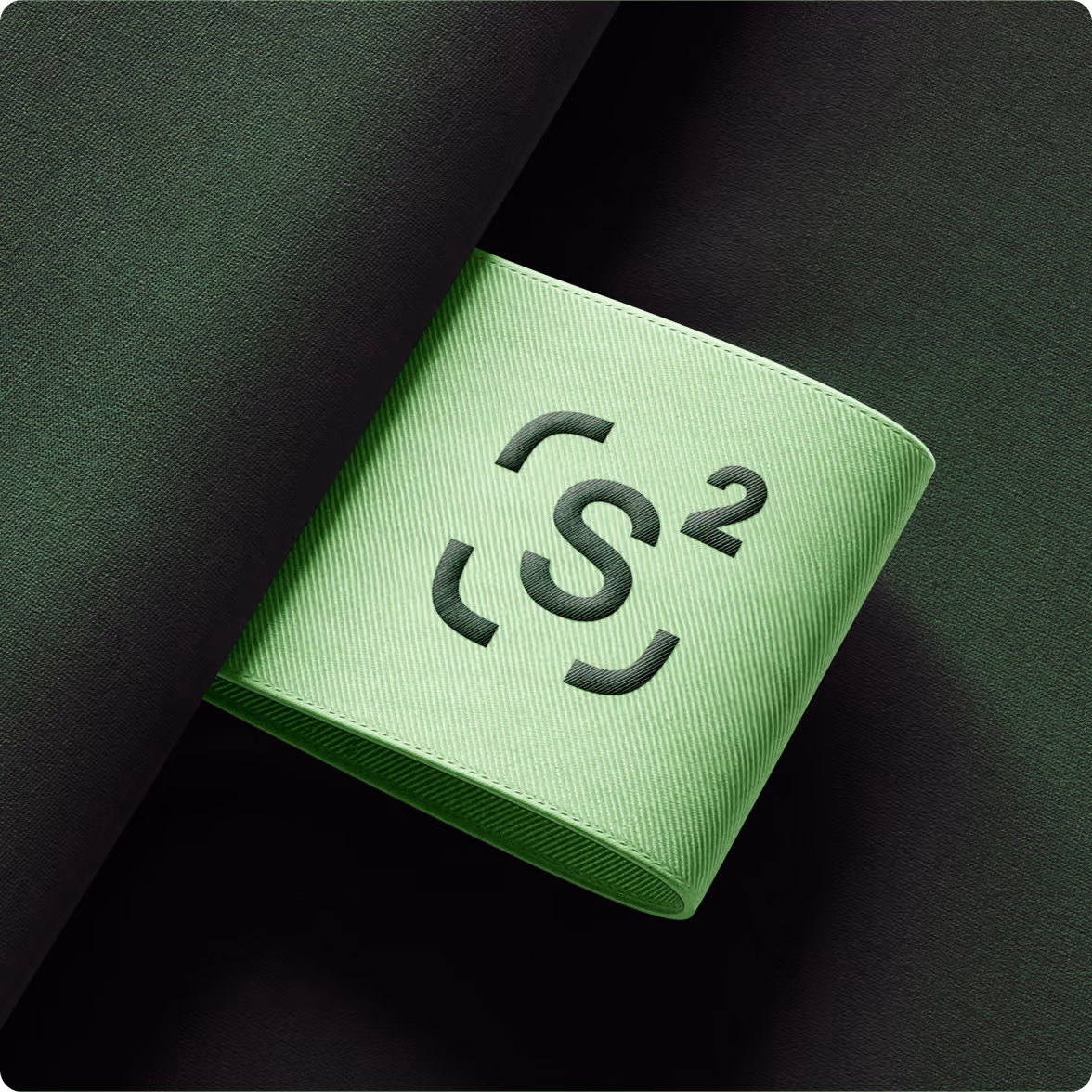 Green woven fabric label with a dark green embroidered design featuring a central 'S' surrounded by four curved segments and a superscript 2, placed against a dark green textured fabric background.