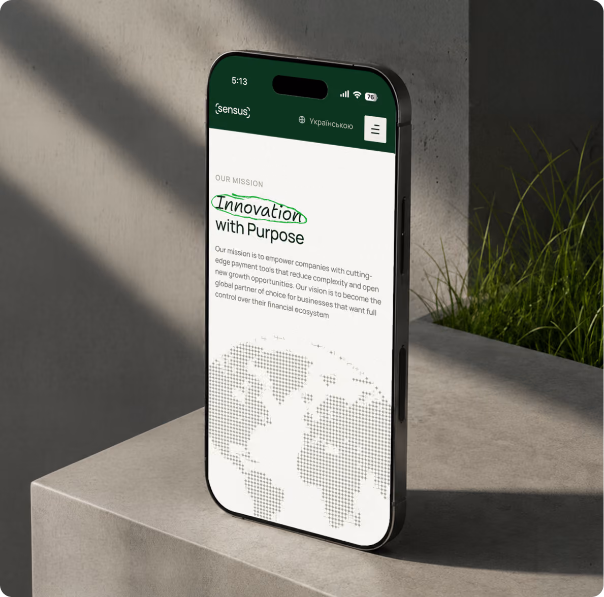 Smartphone standing vertically on a concrete surface displaying a webpage with a green header labeled 'sensus' and text titled 'Innovation with Purpose,' describing a mission to empower companies with cutting-edge payment tools, with a pixelated world map at the bottom of the screen, set against a background of concrete wall and green grass.