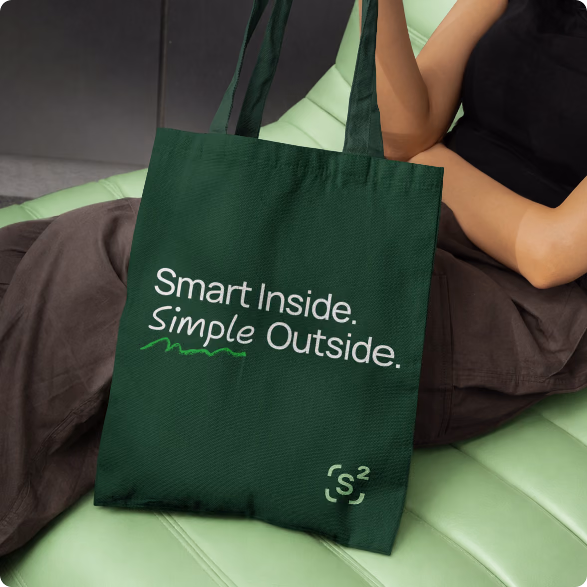 Green tote bag with white text that reads 'Smart Inside. Simple Outside.' and a small s2 logo in the bottom right corner, held by a person sitting on a light green cushioned surface.