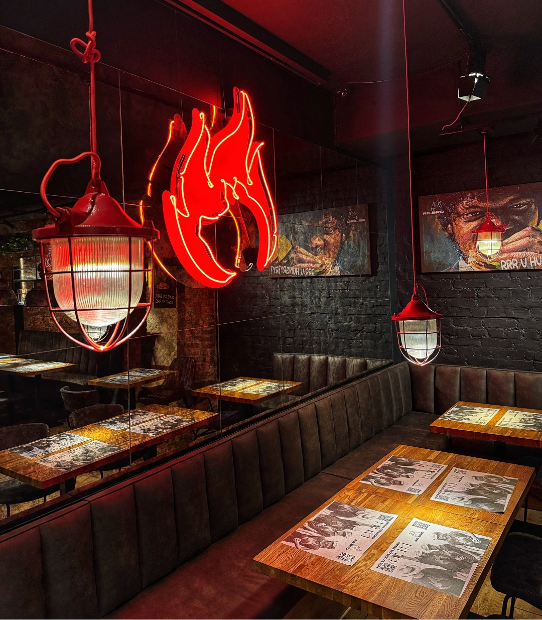 Dark restaurant interior with red hanging lamps, a neon flame sign, wooden tables, and wall art.