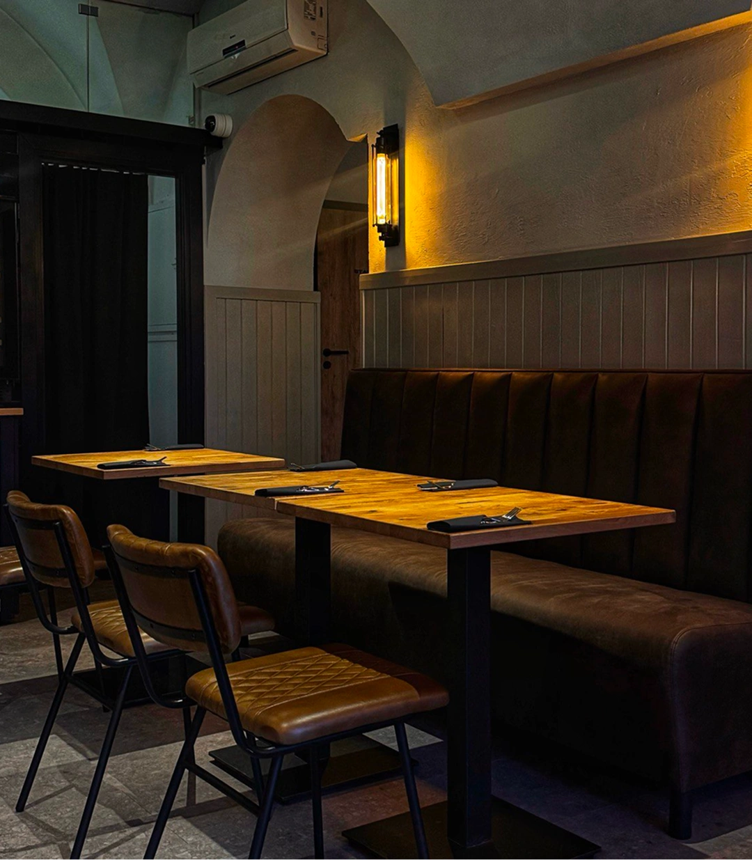 Dimly lit empty restaurant seating with wooden tables, brown leather chairs, and a cushioned bench.