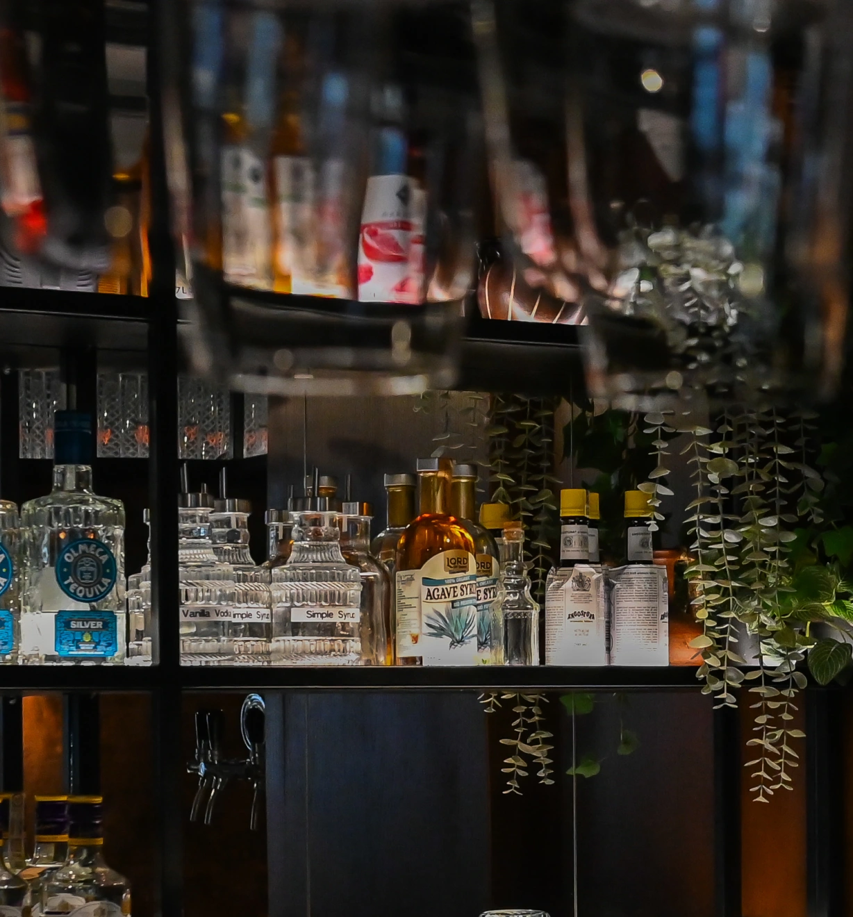 Bar shelf stocked with liquor bottles, glass containers labeled syrup, and hanging greenery.