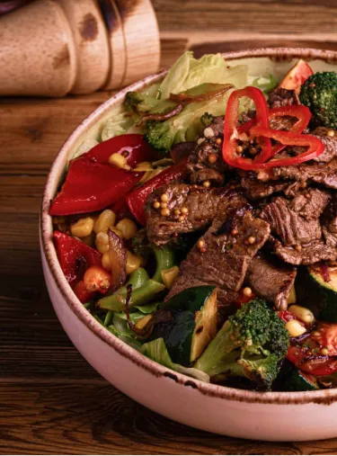 Bowl of grilled beef, corn, broccoli, red bell pepper, and leafy greens on a wooden table.