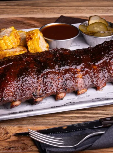 Barbecue ribs glazed with sauce served with corn on the cob, pickles, and a cup of barbecue sauce.