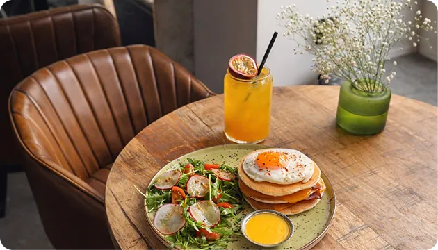 Wooden table with a plate of pancakes topped with fried egg, side salad, sauce, and a glass of orange juice.