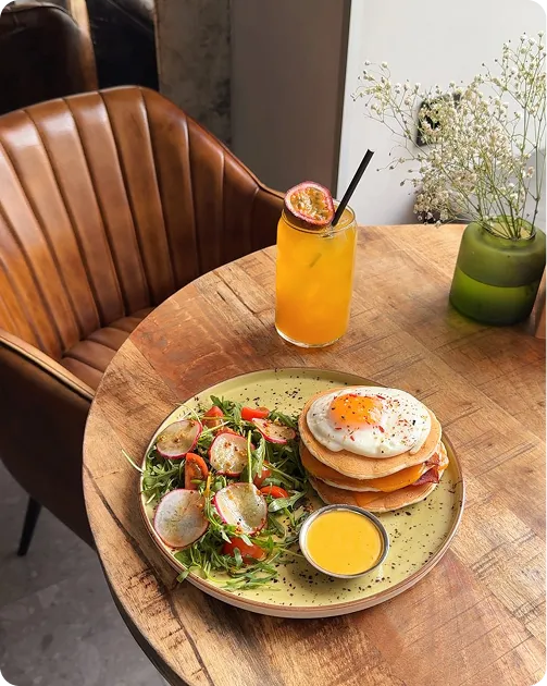 Plate with pancakes topped with a fried egg, salad, and sauce beside a passion fruit drink on a wooden table.