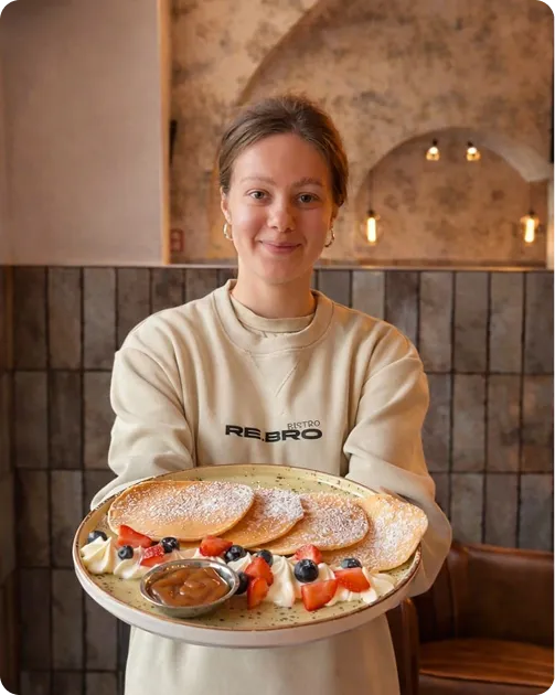 Woman holding plate with three powdered pancakes, fruit, cream, and a small bowl of caramel sauce.