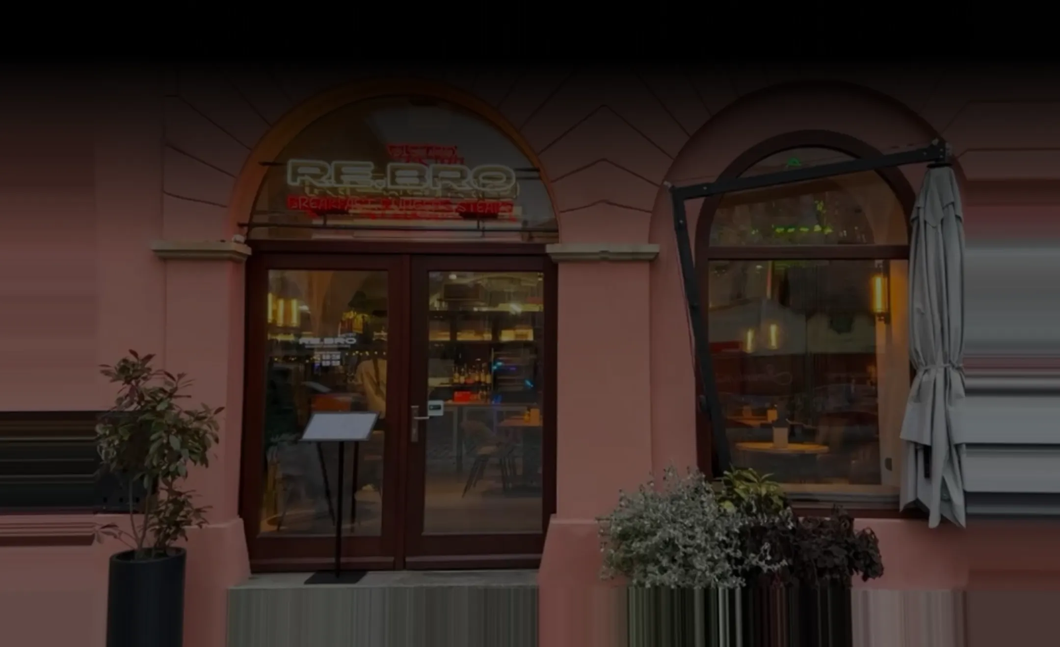 Entrance to a restaurant with a glass door, menu stand, window, plants, and a closed umbrella.