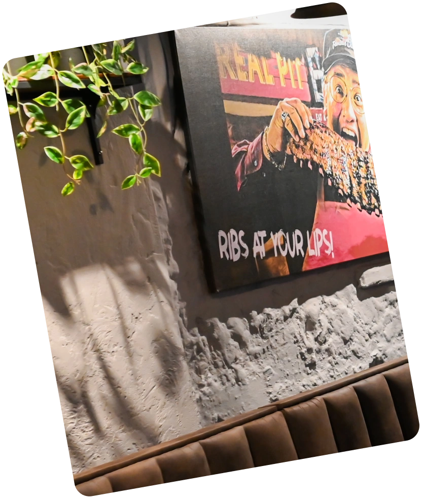 Cozy restaurant corner with brown chairs, wooden tables, wall art, hanging plants, and pendant lights.