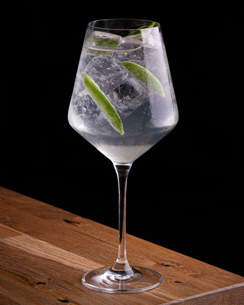 Clear cocktail in a coupe glass garnished with a red flower on a wooden surface against black background.