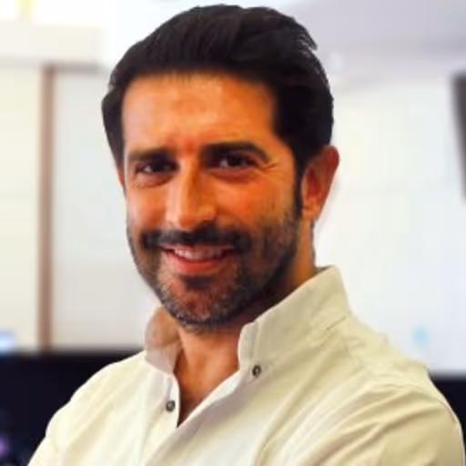 Smiling man with dark hair and beard wearing a white collared shirt indoors.