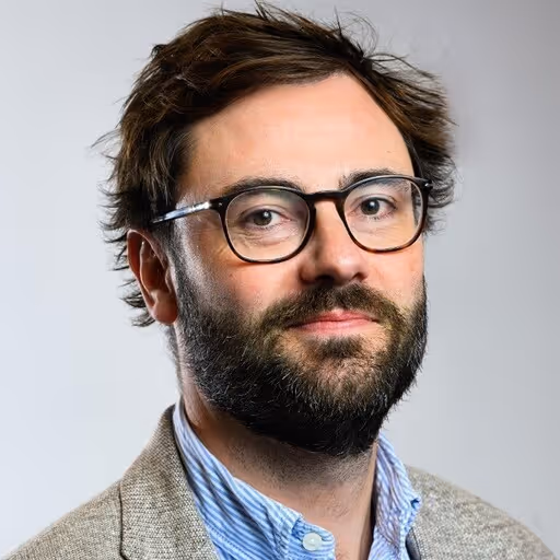 Man with glasses, beard, and brown hair wearing a light blue shirt and gray blazer, looking at camera.