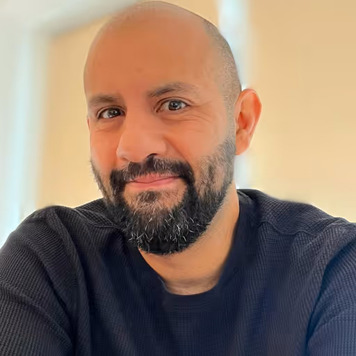 Smiling man with a beard and bald head wearing a dark sweater against a soft background.