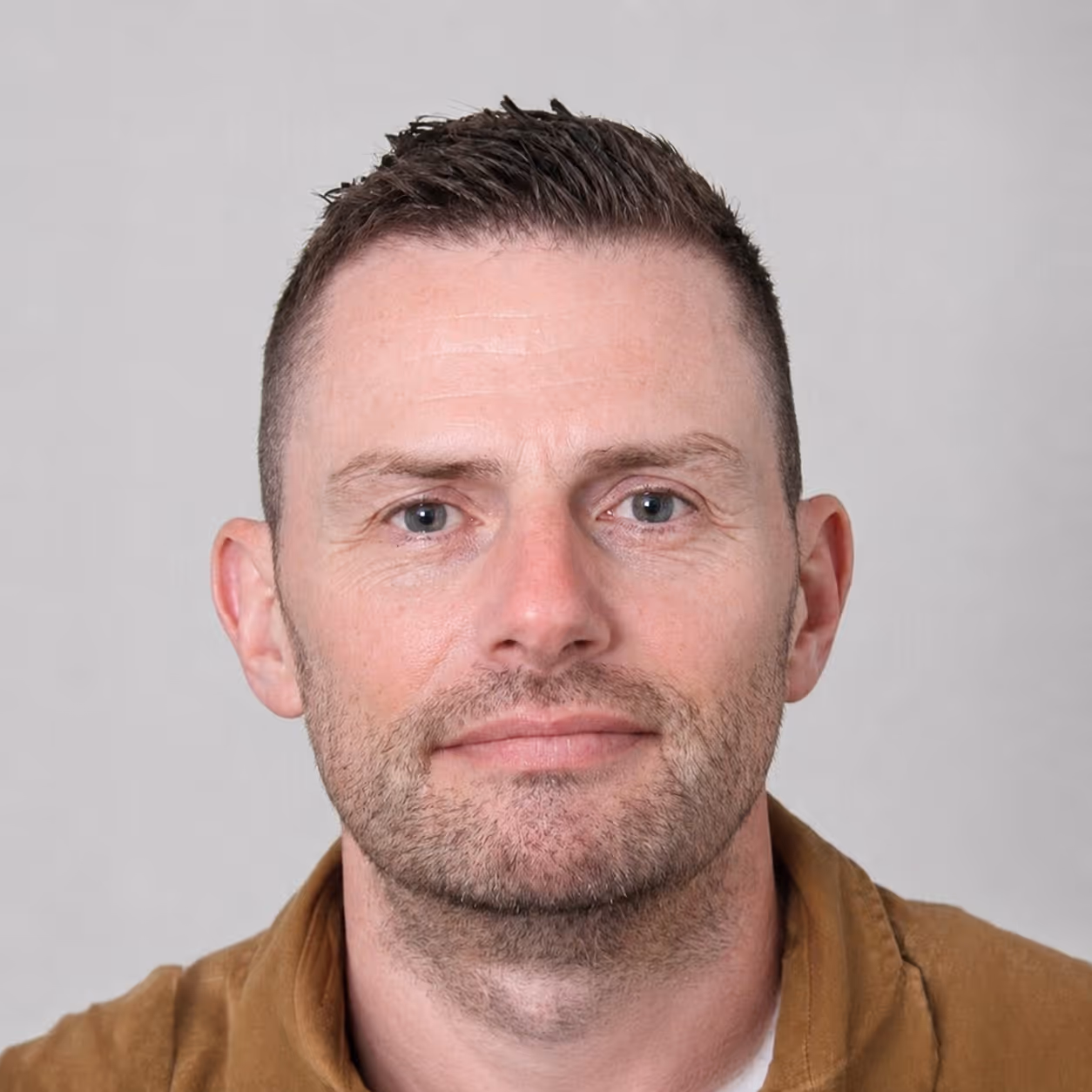 Close-up of a man with short brown hair, light stubble, and blue eyes wearing a brown jacket.