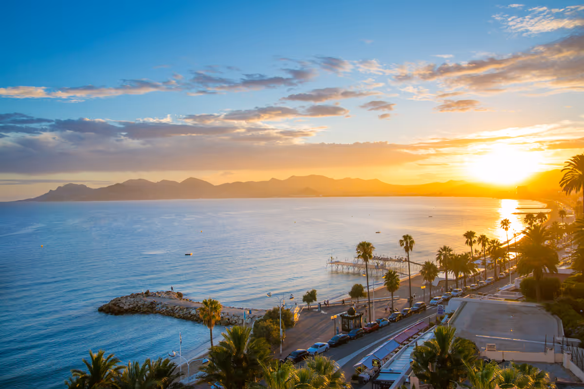 Sunset over a coastal city with palm trees, calm sea, mountains, beach, and parked cars along a road.