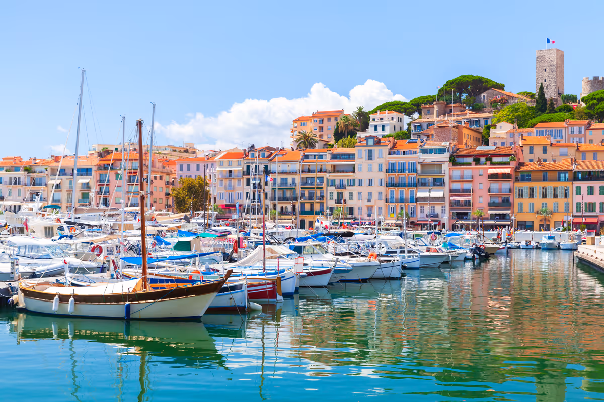 Boats docked in a sunny marina with pastel buildings and a tower flying a French flag in the background.