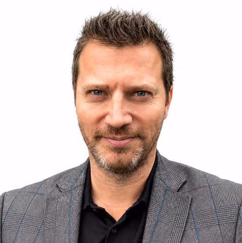 Man with short spiked hair and beard, wearing a gray plaid blazer over a black shirt.