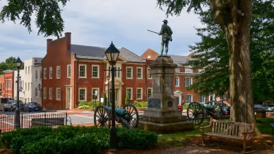 Historic statue of a soldier with cannon artillery on either side, surrounded by trees, a bench, and colonial-style buildings under a cloudy sky.
