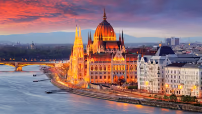 Evening view of the illuminated Hungarian Parliament Building along the Danube River in Budapest with a vibrant sunset sky.