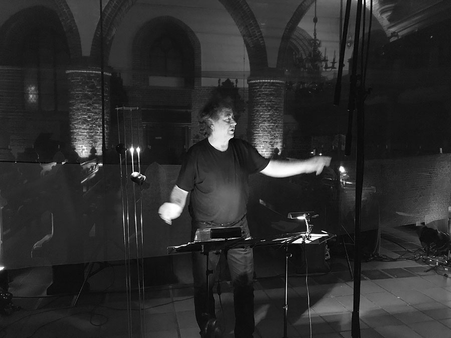 Ein Mann in Schwarz dirigiert in einer Kirche mit gemauerten Bögen und Kerzenlicht.