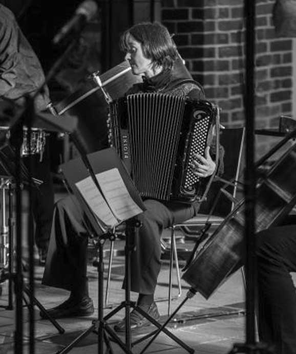 Frau spielt Akkordeon in einem Raum mit Backsteinwand, umgeben von Musikständen und Instrumenten.