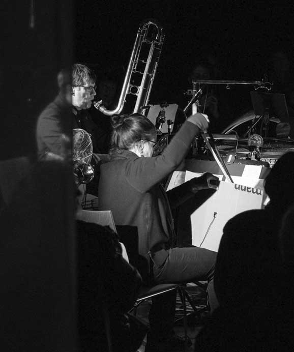 Schwarz-weiß-Bild eines Orchestermusikers, der mit Taktstock dirigiert, und eines Posaunisten im Hintergrund.