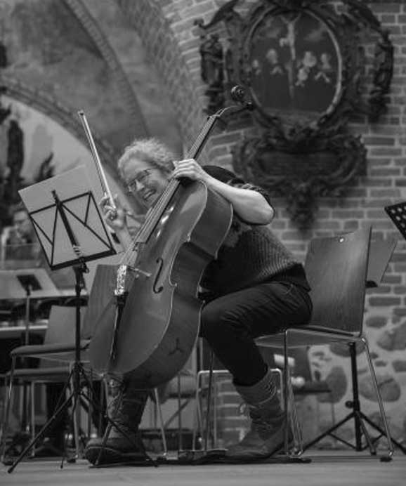 Musikerin sitzt und spielt freudig Cello in einem Raum mit Backsteinwänden und Bilderrahmen.