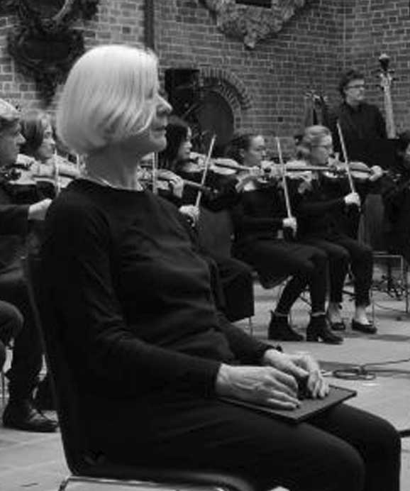 Frau sitzt vor einem Streichorchester mit Violinisten in einem Raum mit Backsteinwänden.