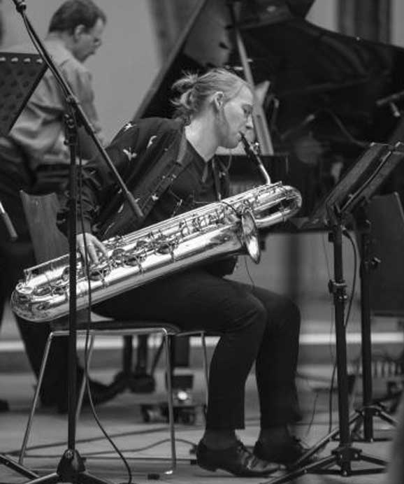 Schwarz-weiß-Foto einer Frau, die ein Baritonsaxophon spielt, daneben ein Klavier.