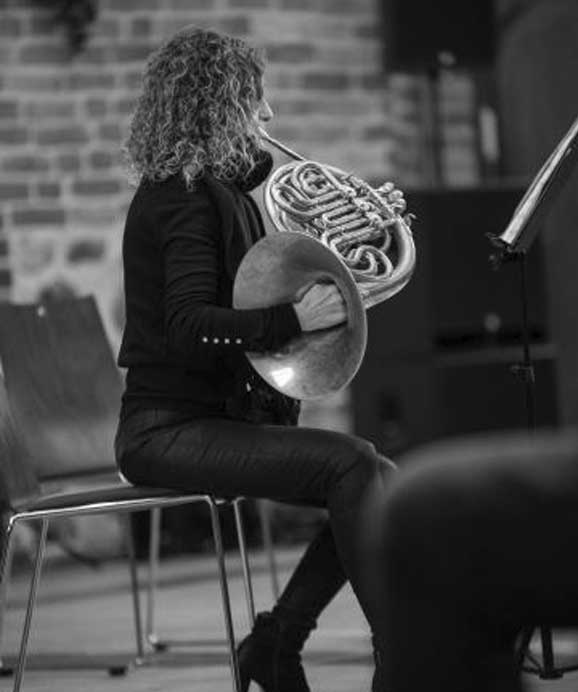Frau mit Locken spielt französisches Horn sitzend vor einem Notenständer in schwarzer Kleidung.